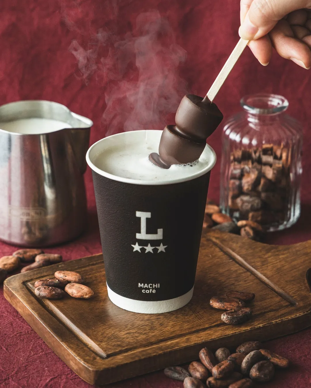 ホットチョコレートとチョコレートのスティックのイメージ写真。白い泡の上にチョコレートスティックが置かれ、湯気が立っている。背景には、ミルクピッチャー、カカオ豆、透明な瓶が置かれている。