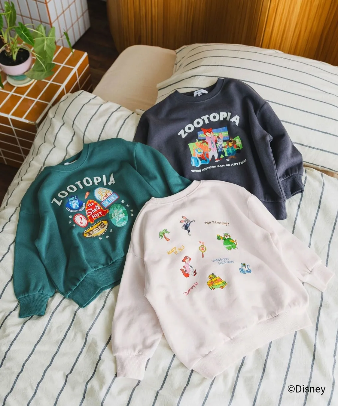 映画『ズートピア』のキャラクターやロゴがプリントされたトレーナーが3枚、布団の上に並べられています。それぞれのトレーナーは異なるデザインで、色も異なります。緑色のトレーナーにはズートピアのロゴと様々な場所のイラストが、グレーのトレーナーにはキャラクター達のイラストと「WHERE ANYONE CAN BE ANYTHING」の文字が、ピンク色のトレーナーにはキャラクター達と「Keep it sly!」などの文字がプリントされています。
