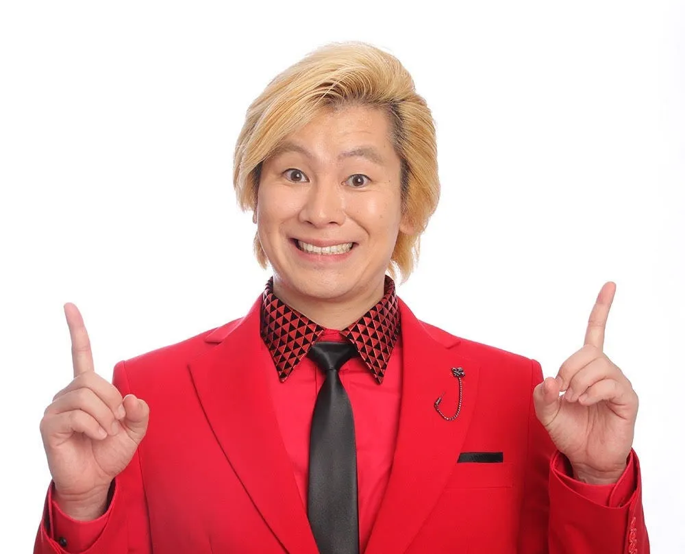 赤いスーツを着用し、笑顔で両手を広げるカズレーザーさんの写真。