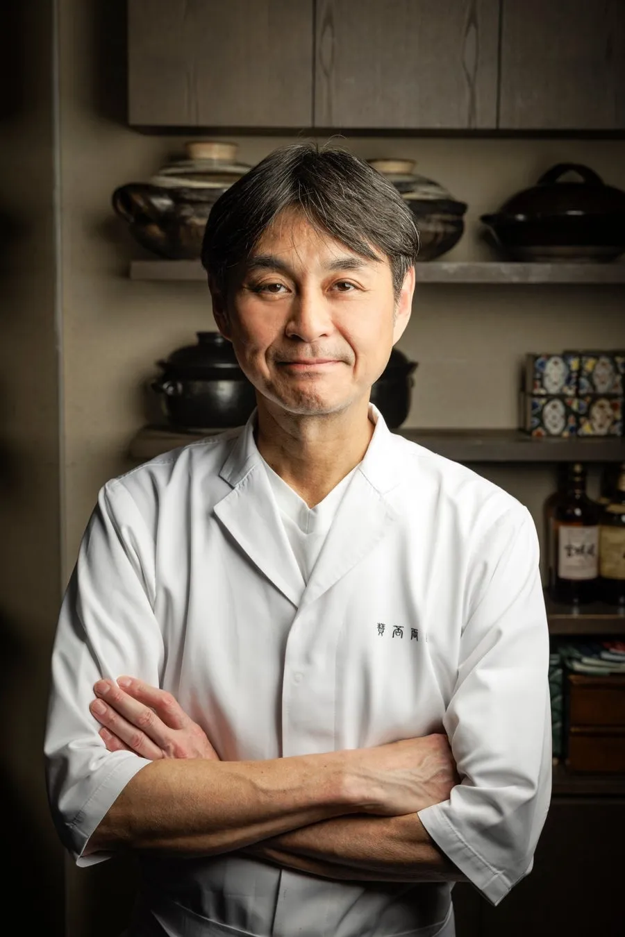 白衣を着た男性が腕を組んで立っている写真。背景には棚があり、陶器や瓶が置かれている。