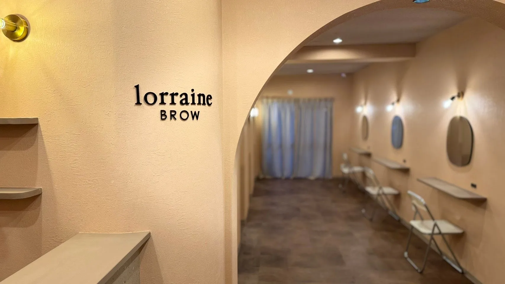 美容サロンの入り口の様子。看板には「lorraine BROW」と書かれており、内装はベージュを基調とした落ち着いた雰囲気です。アーチ型の入り口から奥の空間が見え、鏡や椅子が配置されています。